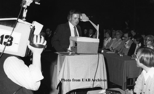 A man stands behind a projector with a new camerman watching